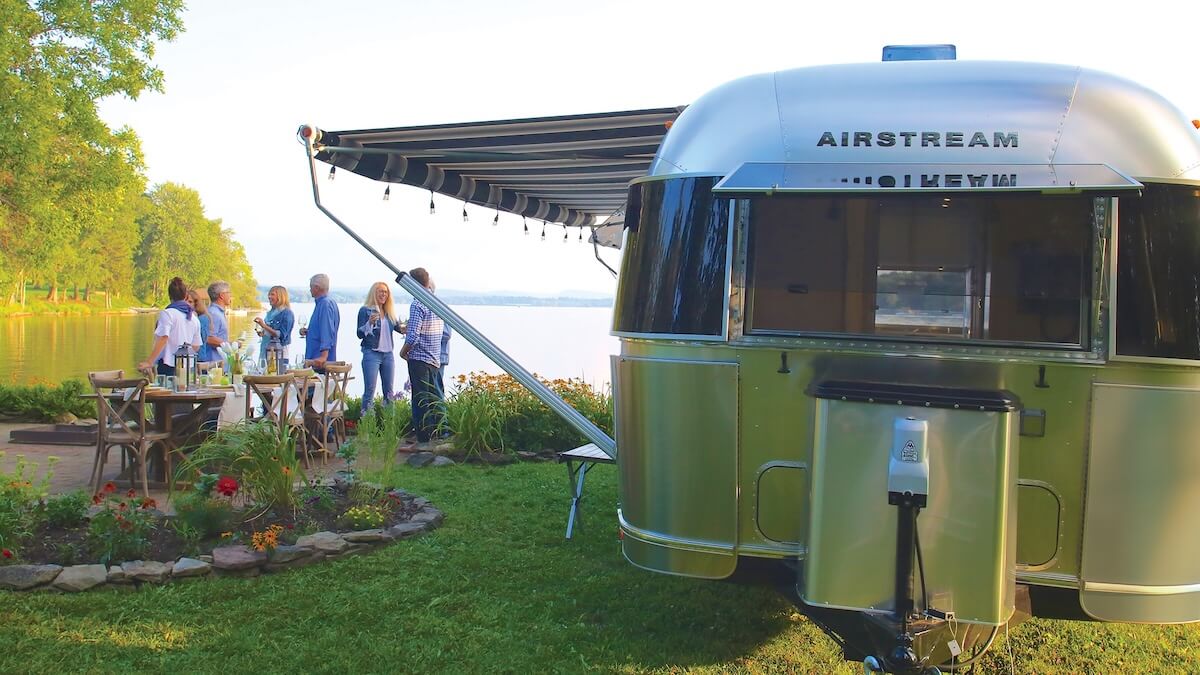 People socializing by an Airstream trailer by a lake
