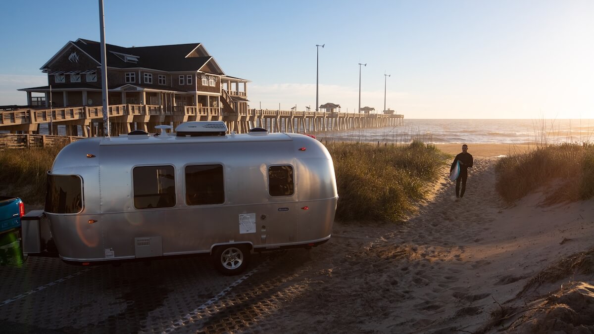 An Airstream trailer parked near the beach with a sufer walking up to it