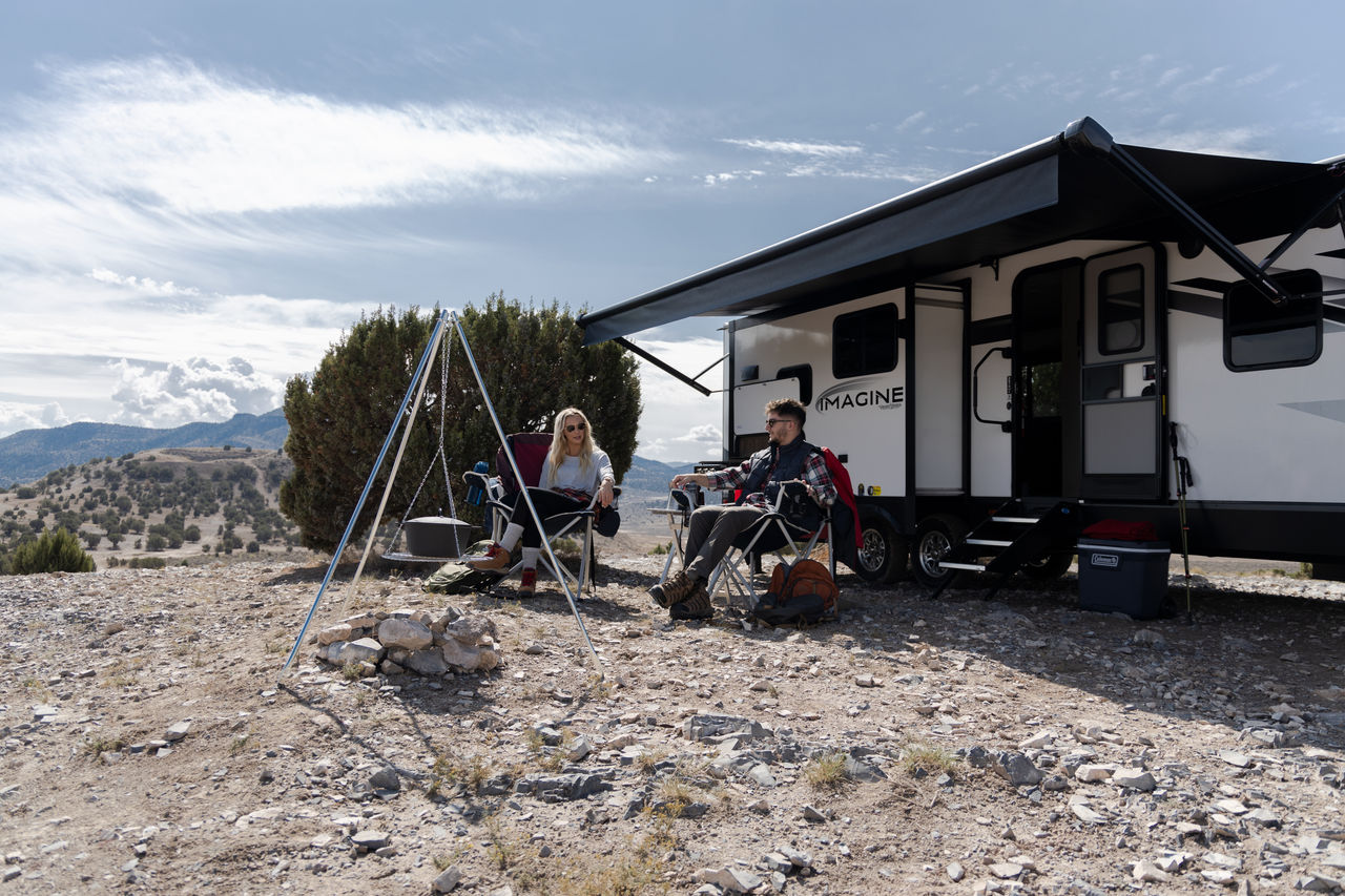 A Grand Design RV, parked in a rocky area with an awning over a couple seated in lawn chairs.